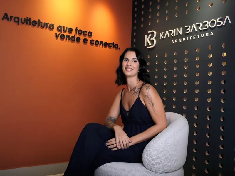 Karin Barbosa sentada em uma poltrona no escritório, entre a parede terracota e a parede navy com a logo